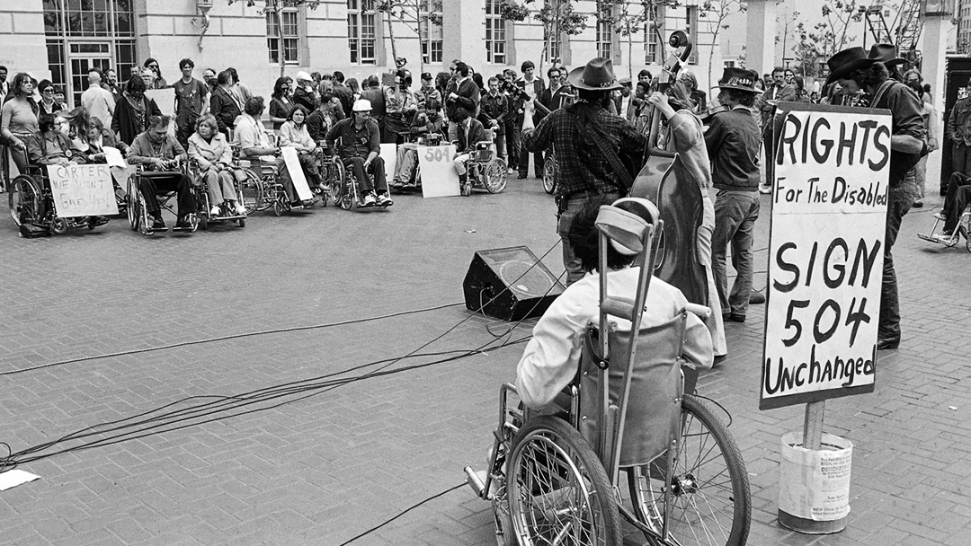 Photograph of a crowd of people in wheelchairs facing a small bad of musicians. A sign that says "Rights for the disabled, sign 504 unchanged"