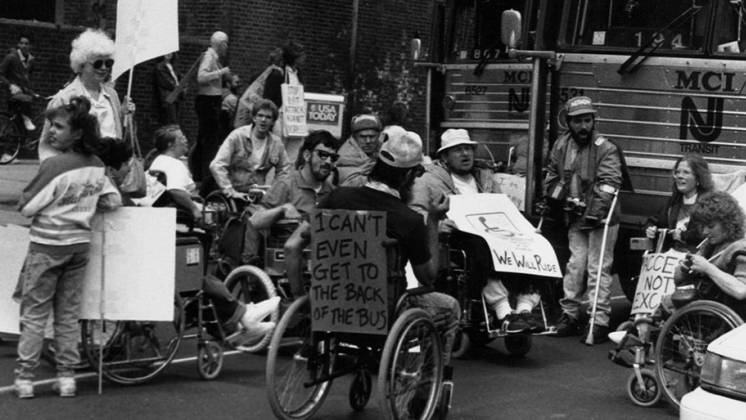 Photograph of a crowd of people, mostly in wheelchairs, in the road in front of a bus. They hold signs, one says "I can't even get to the back of the bus"