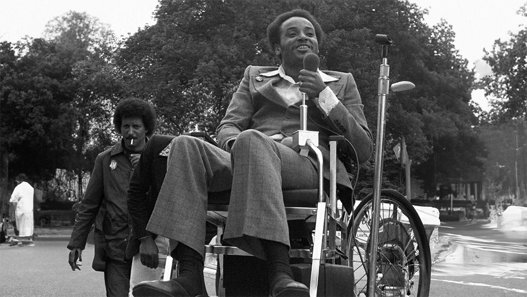 Photograph of a man in a wheelchair, holding a microphone and speaking towards an audience.