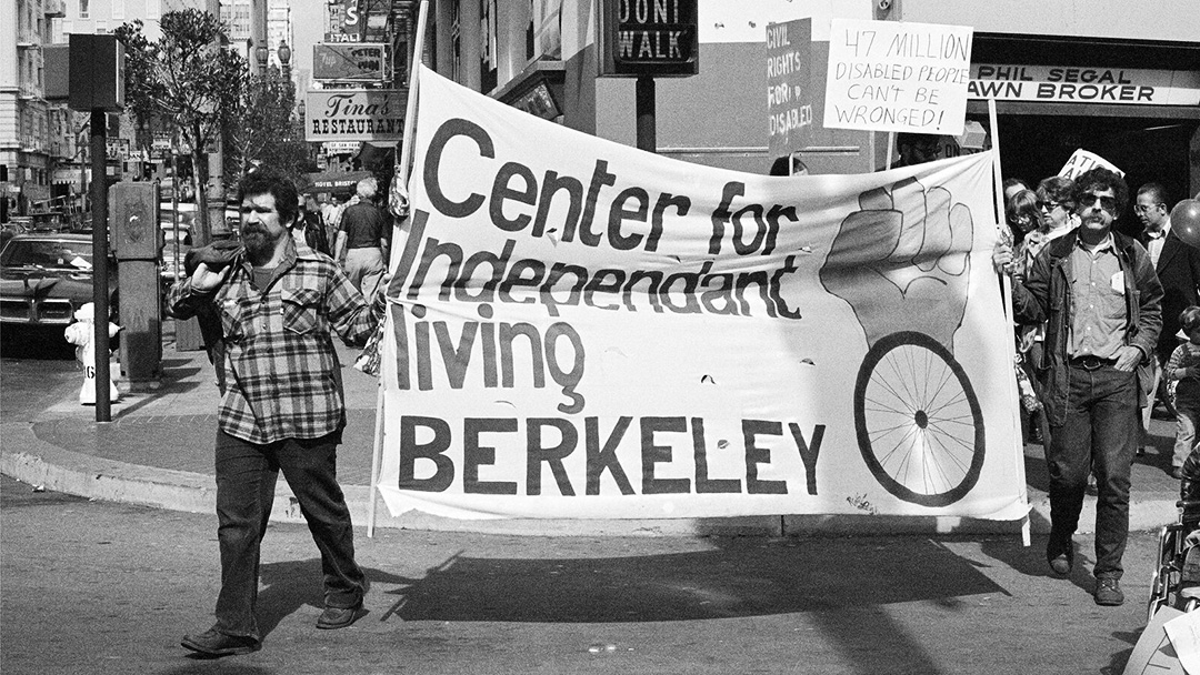 Photograph of two men with mustaches crossing the road, carrying a sign that says "Center for Independent Living Berkeley"