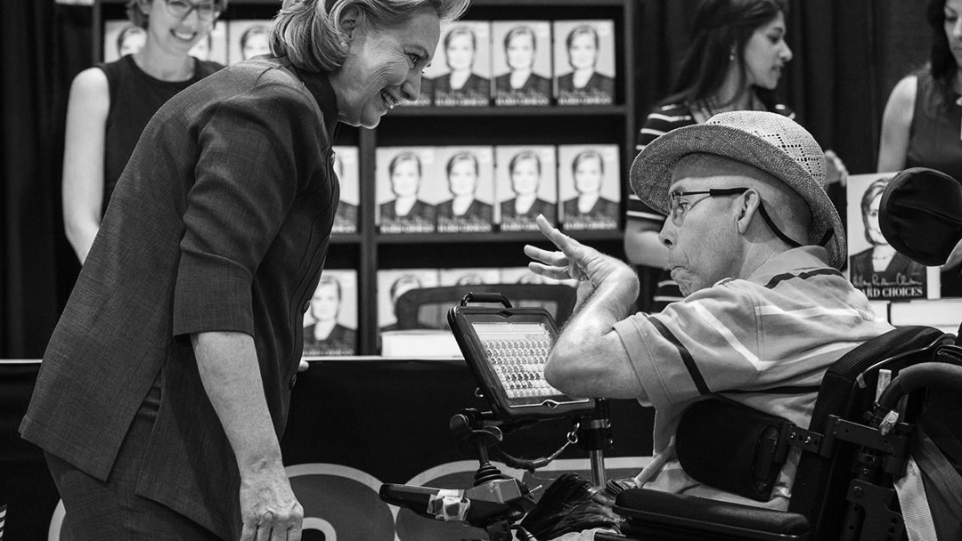 Photograph of Hillary Clinton smiling and leaning towards a man with glasses and a wicker hat in a wheelchair and a iPad.