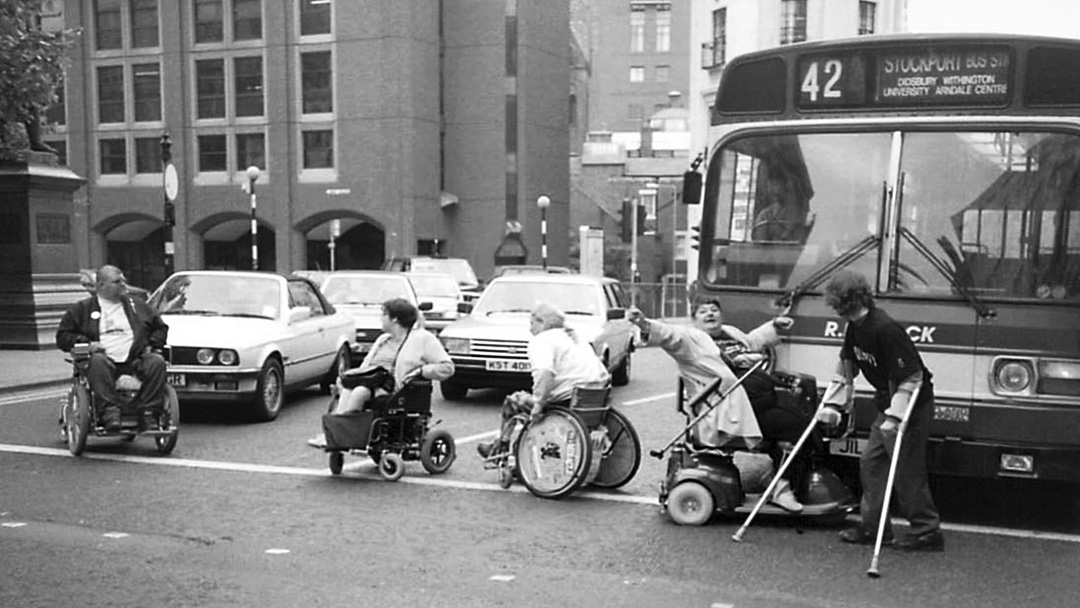 A line of people in wheelchairs, in the street, blocking several cars and a bus.