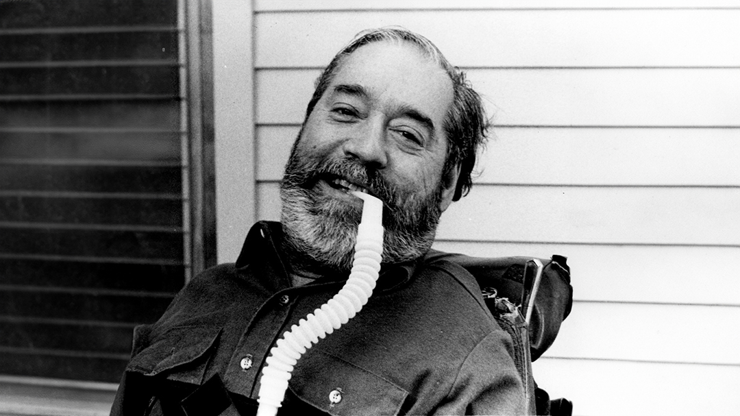 A bearded man leaning back in a wheelchair, smiling, in front of the side of a house with a tube in his mouth.