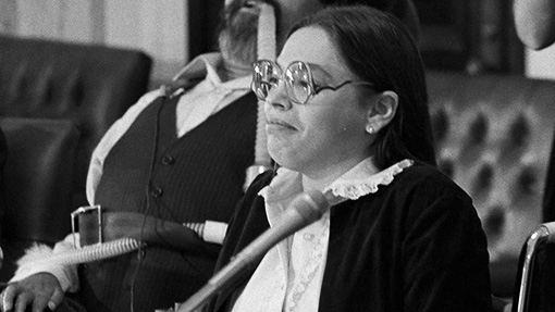 Photograph of a woman with big glasses, smirking, on a panel wearing a button up blouse.