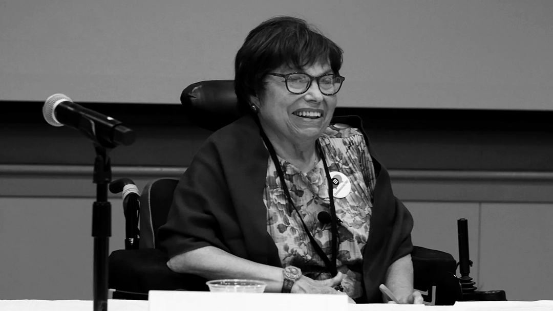 Photograph of a smiling woman in glasses and a wheelchair, behind a microphone, wearing a floral blouse and a sweater.