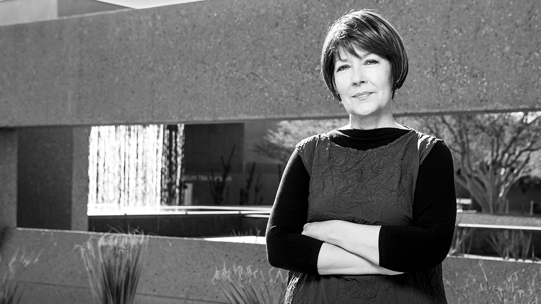 Photograph of a woman with short, round hair in a wrinkled shirt with her arms crossed in front of her, in front of a outdoor architectural installment.